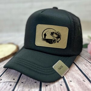 May include: Black trucker hat with a mesh back and a curved peak. The hat features a rectangular patch with a fishing scene: a fisherman, a fish, and a fishing rod. A silver metal tag with the name "TATE" is attached to the peak.