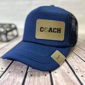May include: A navy blue trucker hat with a black mesh back. The hat features a tan leather patch with the word "COACH" and a basketball graphic. A silver metal tag with the name "MATT" is attached to the brim.