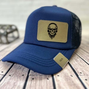 May include: A navy blue trucker cap with a black mesh back and a curved peak. It has a tan leather patch displaying a black skull wearing headphones. A silver clip with the name "GUS" is attached to the brim.
