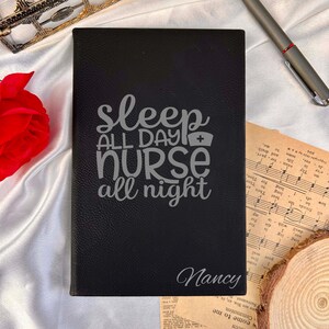 Peut inclure: Journal noir avec le texte "Sleep All Day Nurse All Night" et le nom "Nancy" gravés en argent. Un stylo, une partition et une rose rouge sont visibles en arrière-plan.