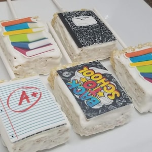 May include: Rice Krispie treats decorated with edible images of school supplies. Treats include images of books, a composition notebook, a paper with an A+, and a "Back to School" graphic. Each treat is on a white stick.