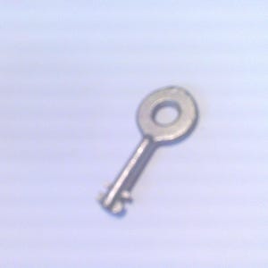 May include: A small, silver-colored key with a circular head and a simple, notched bit. The key is likely made of metal and appears to be a vintage or antique item. The background is a plain, light blue color.