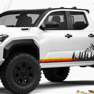 May include: A white pickup truck with black accents and large black tires. The side features a mountain and forest graphic in black, with a horizontal stripe of yellow, red, and orange. The truck has a black roof.