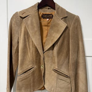 May include: A tan suede blazer featuring a notched lapel, a single front button closure, and two slanted welt pockets. The jacket includes long sleeves with buttoned cuffs, structured shoulders, and a solid brown interior lining with a visible brand label.