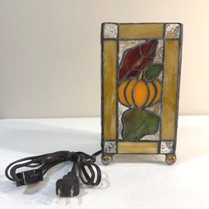 May include: A rectangular stained glass lamp featuring a central orange pumpkin motif surrounded by green and dark red leaves. The lamp has a border of yellow and textured clear glass panels, a black metal frame, and a black electrical cord with a plug.