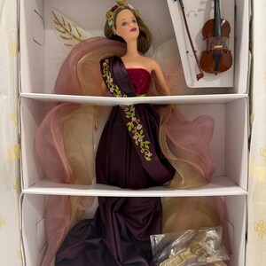 May include: A doll with brown hair wearing a dark purple strapless gown with floral embroidery and sheer pink fabric accents. The doll has a floral headpiece and is packaged with a miniature brown violin, a bow, and a gold-colored doll stand.