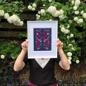 May include: A person is holding a framed print of a colorful, abstract design with pink, blue, and white elements on a dark blue background. The print is framed in white and has a white mat.