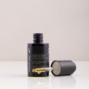 May include: A black glass bottle with a dropper, labeled "future you" and "Youth In Serum". The bottle contains a yellow liquid and is sitting on a white surface.