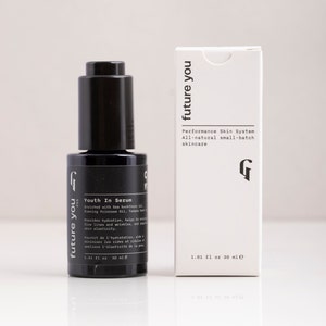 May include: A black glass bottle with a dropper top containing a serum called "Youth In Serum" by the brand "future you". The bottle is labeled with the text "Enriched with Sea buckthorn oil, Evening Primrose Oil, Tamanu Seed Oil, Rose Oil" and "Provides hydration, helps to minimize fine lines and wrinkles, and improve skin elasticity". The bottle is sitting on a white surface next to a white box with the same branding and text. The box is labeled with the text "Performance Skin System, All-natural small-batch skincare".
