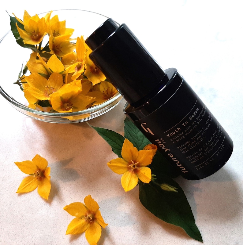 May include: A black glass bottle with a dropper top and a white label that reads "future you PSS Youth In Serum" with a description of the serum's benefits. The bottle is sitting on a white surface with yellow flowers.