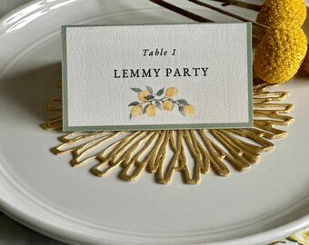 May include: A rectangular white paper place card with a sage green border, featuring the black text Table 1 and LEMMY PARTY above a watercolor illustration of yellow lemons on a branch, resting on a gold-colored circular decorative mat.
