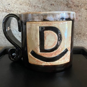 Pode incluir: Uma caneca de cerâmica com esmalte castanho escuro brilhante e borda branca. Uma placa quadrada bege está centrada na frente, apresentando uma letra D em relevo acima de uma linha curva. A caneca tem uma alça grande e curva.