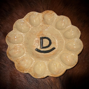 May include: A round, beige ceramic egg serving platter with a scalloped edge and twelve oval indentations arranged in a circle. The center of the platter features a large, dark, hand-painted letter D with a curved line underneath it.
