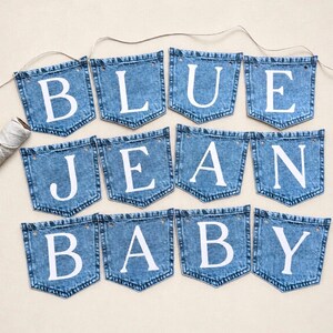 May include: A decorative banner consisting of twelve blue denim pockets arranged in three rows, each featuring a single white letter. The letters spell out the phrase BLUE JEAN BABY. A thin, light-colored string connects the pockets at the top.