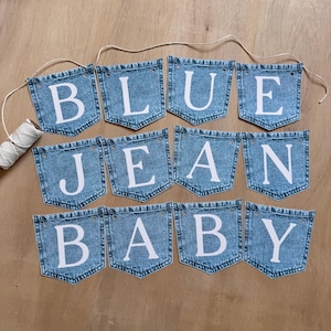 May include: A decorative banner consisting of twelve individual denim pocket-shaped fabric pieces strung together on a white cord. Each pocket features a large white capital letter, collectively spelling out the phrase BLUE JEAN BABY.