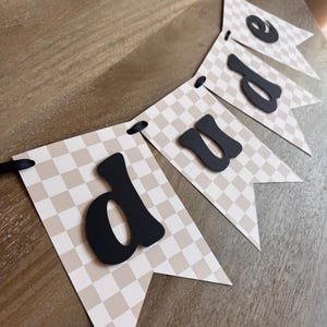 May include: A banner with pennant flags in a beige and white checkered pattern. Black letters spell out the word "dude". The banner is strung with a black ribbon and is displayed on a wooden surface.