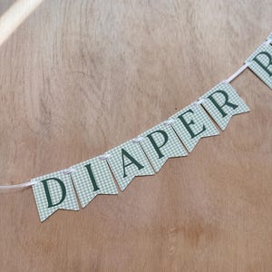 K&ouml;nnte beinhalten: Ein Banner mit dem Wort "DIAPER" in dunkelgr&uuml;nen Buchstaben. Die Wimpel haben ein hellgr&uuml;nes und wei&szlig;es Karomuster. Das Banner ist an einem wei&szlig;en Band auf einem Holzgrund befestigt.