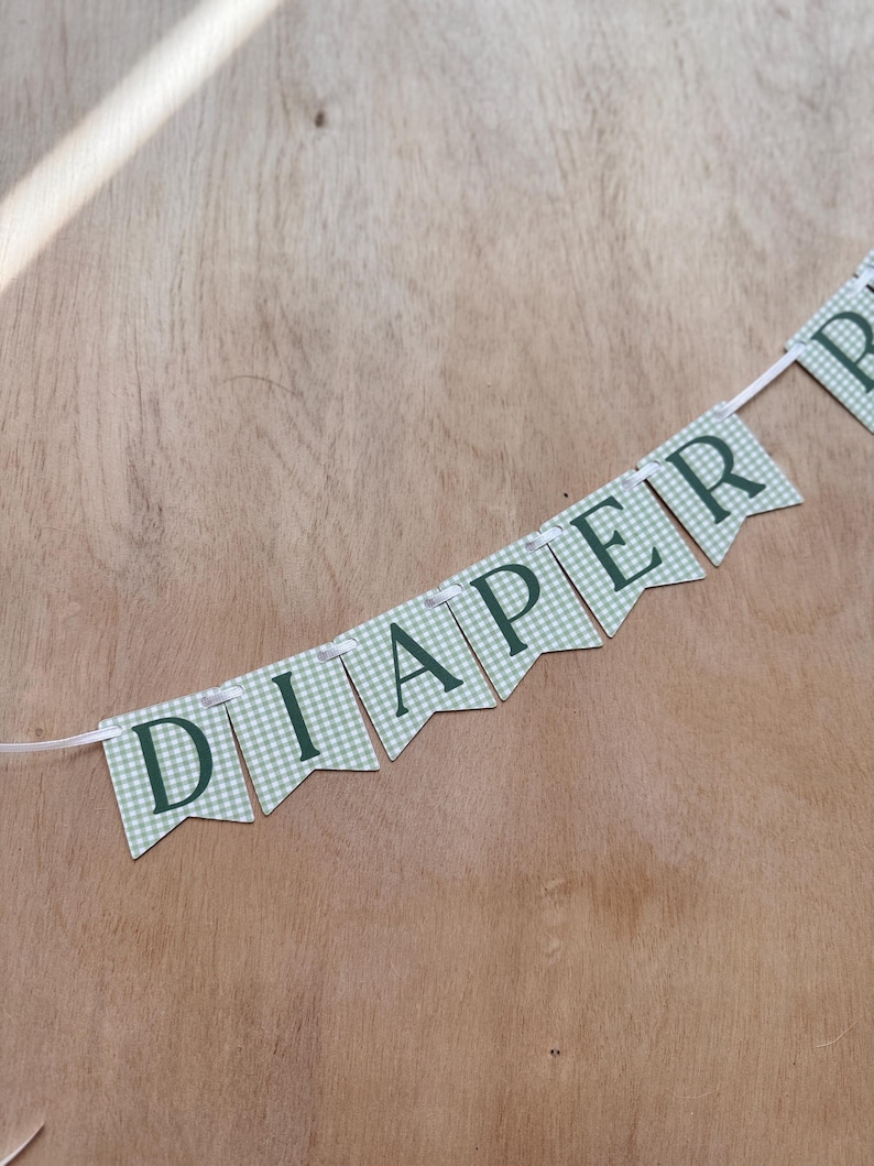 K&ouml;nnte beinhalten: Ein Banner mit dem Wort "DIAPER" in dunkelgr&uuml;nen Buchstaben. Die Wimpel haben ein hellgr&uuml;nes und wei&szlig;es Karomuster. Das Banner ist an einem wei&szlig;en Band auf einem Holzgrund befestigt.