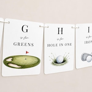 May include: A white paper banner featuring golf-themed alphabet cards. One card shows a green with a red flag and the text G is for GREENS. Another card displays a golf ball splashing into a hole with the text H is for HOLE IN ONE.