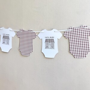 May include: A garland of baby onesies in white and brown. Two white onesies feature a black and white cafe illustration and the text "CAFE BEBE". The other two onesies have a brown and white checkered pattern.