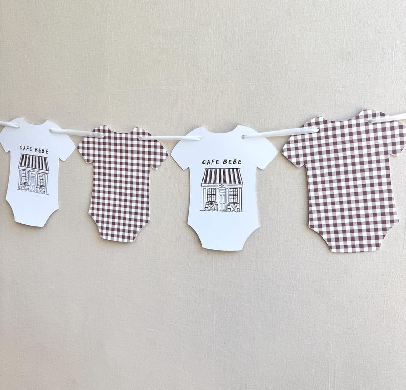 May include: A garland of baby onesies in white and brown. Two white onesies feature a black and white cafe illustration and the text "CAFE BEBE". The other two onesies have a brown and white checkered pattern.