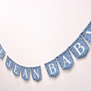 May include: A decorative banner featuring twelve individual blue denim pockets strung together on a white cord. Each pocket displays a single white capital letter, collectively spelling out the phrase BLUE JEAN BABY in a clear, sans-serif font.