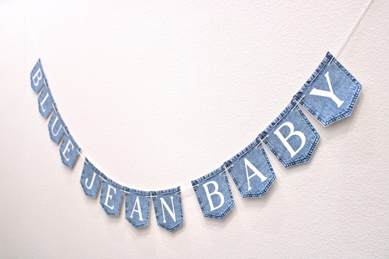 May include: A decorative banner featuring twelve individual blue denim pockets strung together on a white cord. Each pocket displays a single white capital letter, collectively spelling out the phrase BLUE JEAN BABY in a clear, sans-serif font.