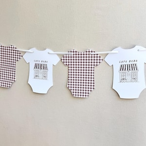 May include: A garland of baby onesies. Two onesies have a brown and white gingham pattern. The other two are white with a black and white illustration of a cafe and the text "CAFE BEBE". The garland is strung on a white string.