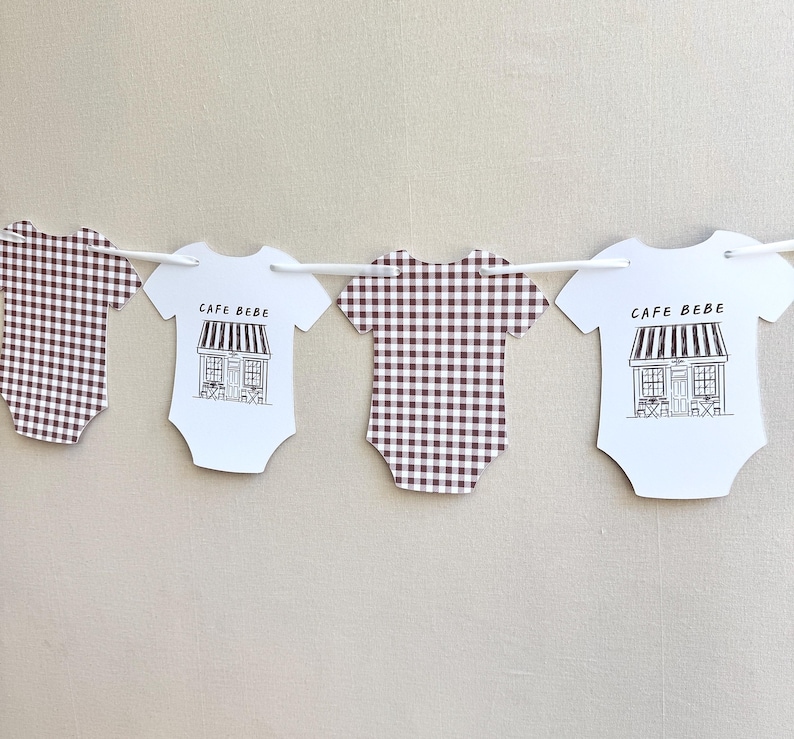 May include: A garland of baby onesies. Two onesies have a brown and white gingham pattern. The other two are white with a black and white illustration of a cafe and the text "CAFE BEBE". The garland is strung on a white string.
