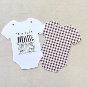 May include: Two baby bodysuit-shaped paper cutouts. One is white with a black and white cafe illustration and the text "CAFE BEBE". The other is brown and white gingham.