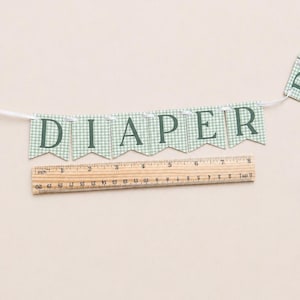 K&ouml;nnte beinhalten: Ein Banner mit dem Wort "DIAPER" in dunkelgr&uuml;nen Buchstaben. Jeder Wimpel hat ein hellgr&uuml;nes und wei&szlig;es Vichy-Muster. Ein Holzlineal mit Zentimetermessungen befindet sich darunter.