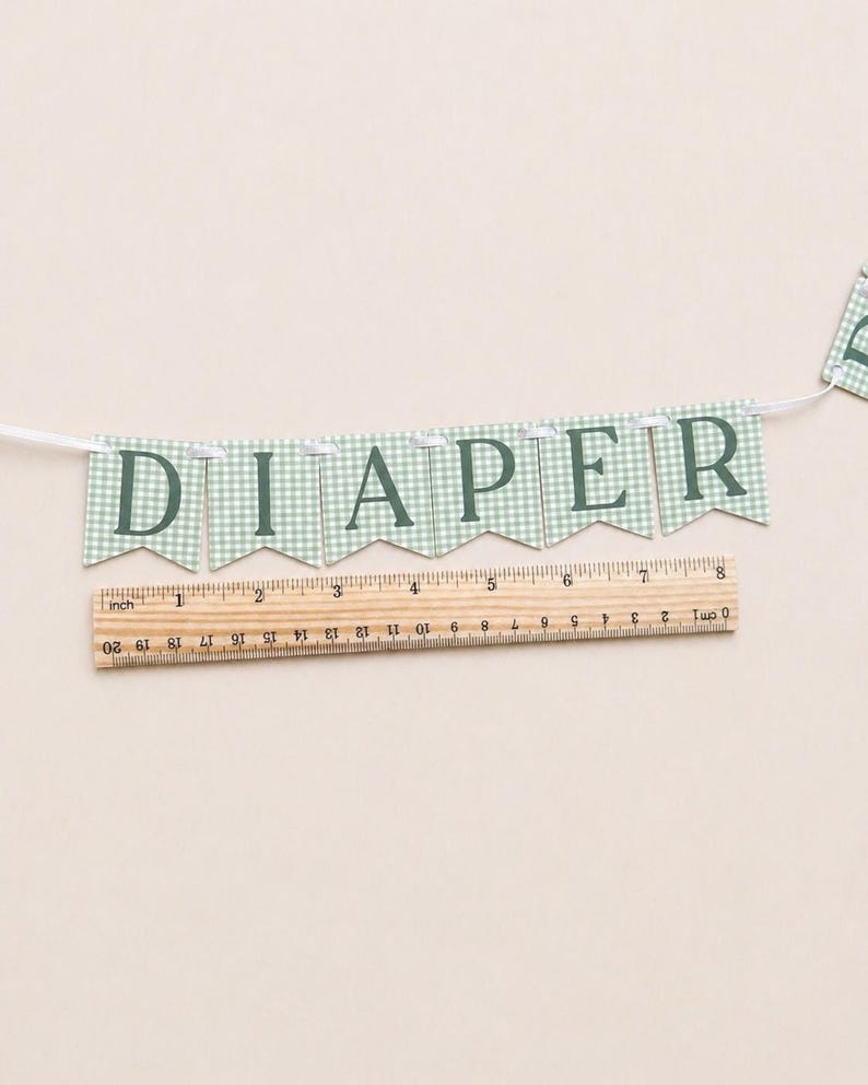 K&ouml;nnte beinhalten: Ein Banner mit dem Wort "DIAPER" in dunkelgr&uuml;nen Buchstaben. Jeder Wimpel hat ein hellgr&uuml;nes und wei&szlig;es Vichy-Muster. Ein Holzlineal mit Zentimetermessungen befindet sich darunter.