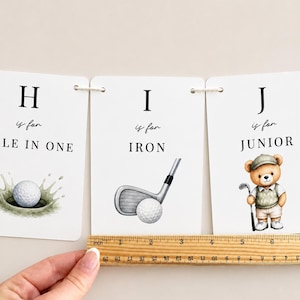 May include: Three white rectangular cards featuring letters H, I, and J with golf-themed illustrations and text. A wooden ruler is placed along the bottom edge, showing a width of approximately 15 cm for the cards combined.