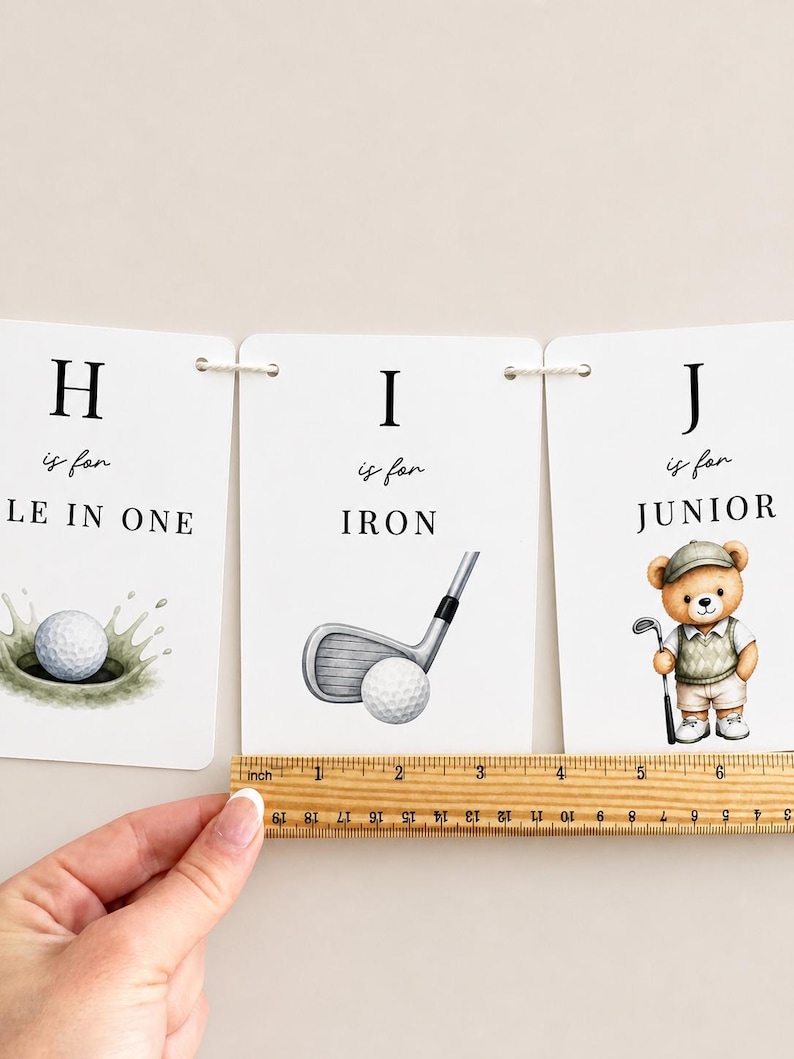 May include: Three white rectangular cards featuring letters H, I, and J with golf-themed illustrations and text. A wooden ruler is placed along the bottom edge, showing a width of approximately 15 cm for the cards combined.