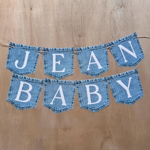May include: A decorative banner featuring eight individual denim-style pockets strung together on a thin cord. Each pocket is printed with a single white capital letter, collectively spelling out the phrase JEAN BABY in two rows of four pockets.