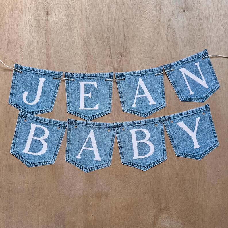 May include: A decorative banner featuring eight individual denim-style pockets strung together on a thin cord. Each pocket is printed with a single white capital letter, collectively spelling out the phrase JEAN BABY in two rows of four pockets.