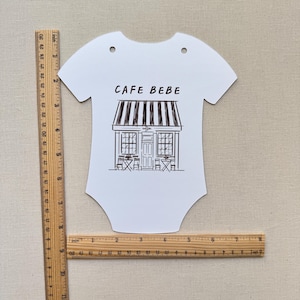 May include: White baby bodysuit-shaped sign with the text "CAFE BEBE" above a black and white illustration of a cafe. The sign has two holes at the top and is approximately 10 inches long.