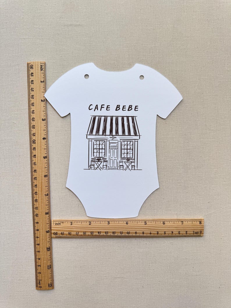May include: White baby bodysuit-shaped sign with the text "CAFE BEBE" above a black and white illustration of a cafe. The sign has two holes at the top and is approximately 10 inches long.