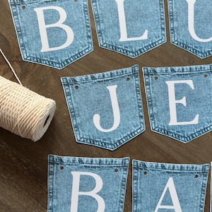 May include: A decorative banner consisting of multiple blue denim pocket-shaped pennants, each featuring a large white letter. The letters spell out BLUE JEANS BABY. Each pennant has two holes at the top for stringing, accompanied by a spool of white twine.