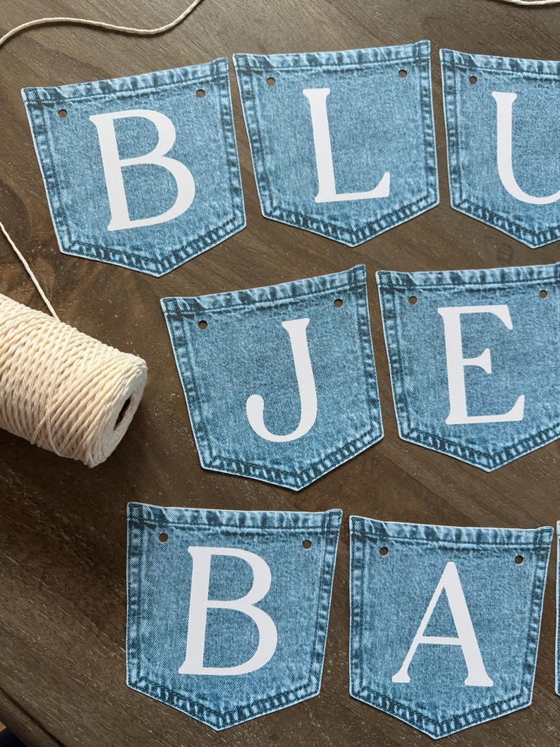 May include: A decorative banner consisting of multiple blue denim pocket-shaped pennants, each featuring a large white letter. The letters spell out BLUE JEANS BABY. Each pennant has two holes at the top for stringing, accompanied by a spool of white twine.