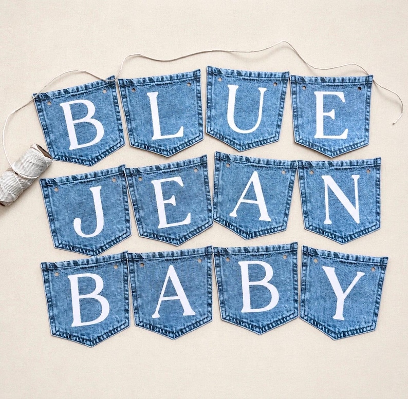 May include: A decorative banner consisting of twelve blue denim pockets arranged in three rows, each featuring a single white letter. The letters spell out the phrase BLUE JEAN BABY. A thin, light-colored string connects the pockets at the top.