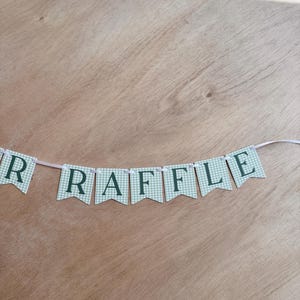 K&ouml;nnte beinhalten: Ein Banner mit dem Wort "RAFFLE" in dunkelgr&uuml;nen Buchstaben auf hellgr&uuml;nen und wei&szlig;en karierten Wimpeln. Das Banner ist an einem d&uuml;nnen wei&szlig;en Band auf einem Holzgrund befestigt.