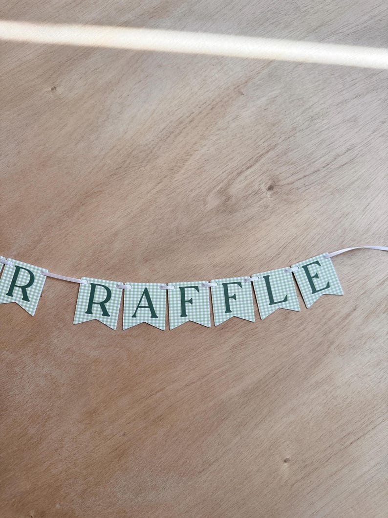 K&ouml;nnte beinhalten: Ein Banner mit dem Wort "RAFFLE" in dunkelgr&uuml;nen Buchstaben auf hellgr&uuml;nen und wei&szlig;en karierten Wimpeln. Das Banner ist an einem d&uuml;nnen wei&szlig;en Band auf einem Holzgrund befestigt.