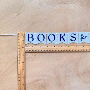 K&ouml;nnte beinhalten: Ein Banner mit der Aufschrift "BOOKS for" in marineblauen Buchstaben. Das Banner besteht aus blau-wei&szlig; karierten Flaggen. Ein Holzlineal liegt daneben und misst in Zoll. Das Lineal ist etwa 50 cm lang.