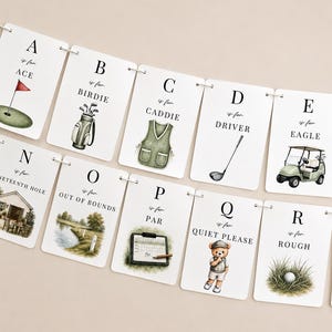May include: A decorative banner featuring white rectangular cards strung together, each displaying a letter of the alphabet from A to R with golf-themed illustrations and text such as A is for ACE, B is for BIRDIE, C is for CADDIE, D is for DRIVER, E is for EAGLE, N is for NINETEENTH HOLE, O is for OUT OF BOUNDS, P is for PAR, Q is for QUIET PLEASE, and R is for ROUGH.