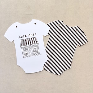 May include: Two baby bodysuit-shaped paper cutouts. One is white with a black and white cafe illustration and the text "CAFE BEBE". The other is a black and white striped pattern.