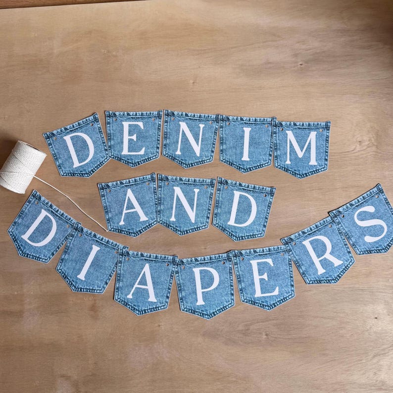 May include: A decorative banner composed of individual blue denim pocket-shaped fabric pieces, each featuring a large white letter. The letters spell out the phrase DENIM AND DIAPERS. A spool of white string is positioned to the left of the banner.