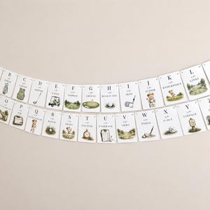 May include: A decorative banner featuring twenty-six white rectangular cards arranged in alphabetical order from A to Z. Each card displays a letter, a golf-themed word, and a watercolour illustration of golf equipment or a bear playing golf.
