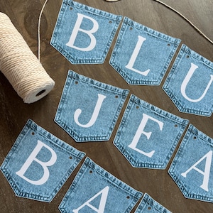 May include: A banner made of blue denim pockets arranged in two rows, each featuring a large white letter. The letters spell out BLUE JEANS BABY. A spool of white twine is positioned to the left, with a length of twine threaded through the top holes.