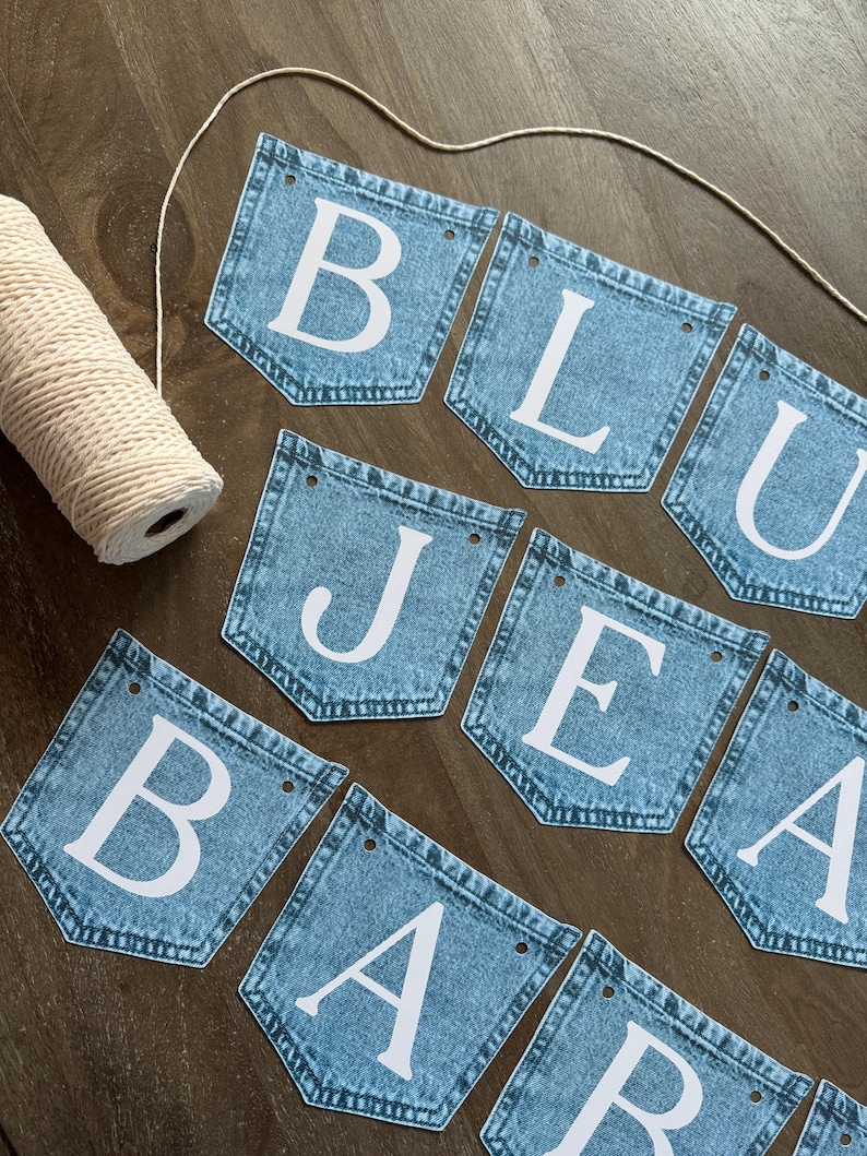 May include: A banner made of blue denim pockets arranged in two rows, each featuring a large white letter. The letters spell out BLUE JEANS BABY. A spool of white twine is positioned to the left, with a length of twine threaded through the top holes.